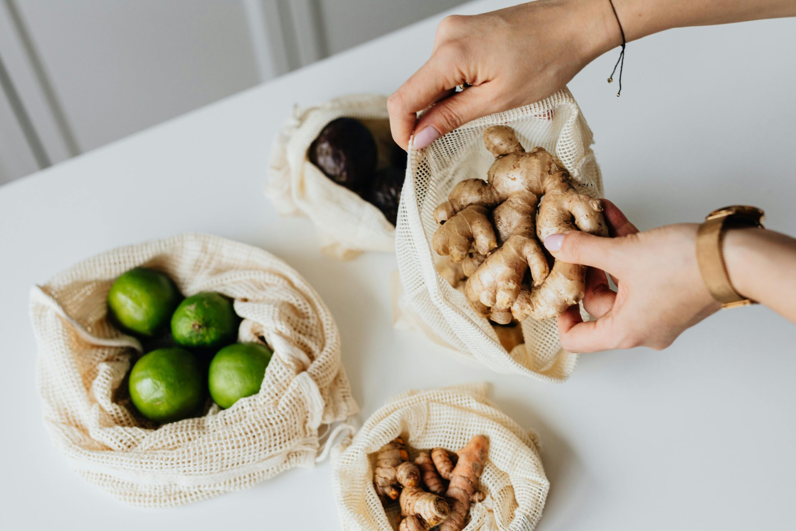 Hands sorting ginger, limes, avocados, and turmeric in reusable net bags on a white surface.